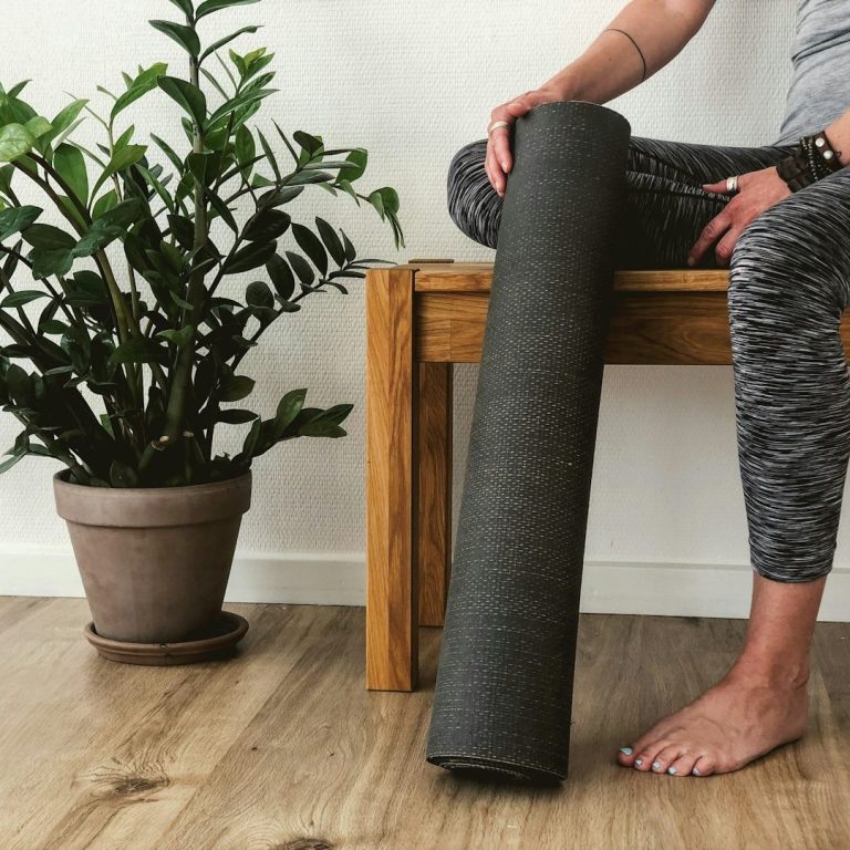 Frau in sportlicher Kleidung sitzt mit ihrer Yoga Matte in der Hand auf einer Bank